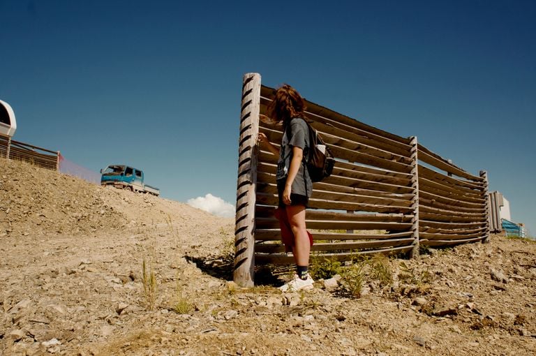 A woman stands by a wooden fence on a sunny day, with a truck in the background.