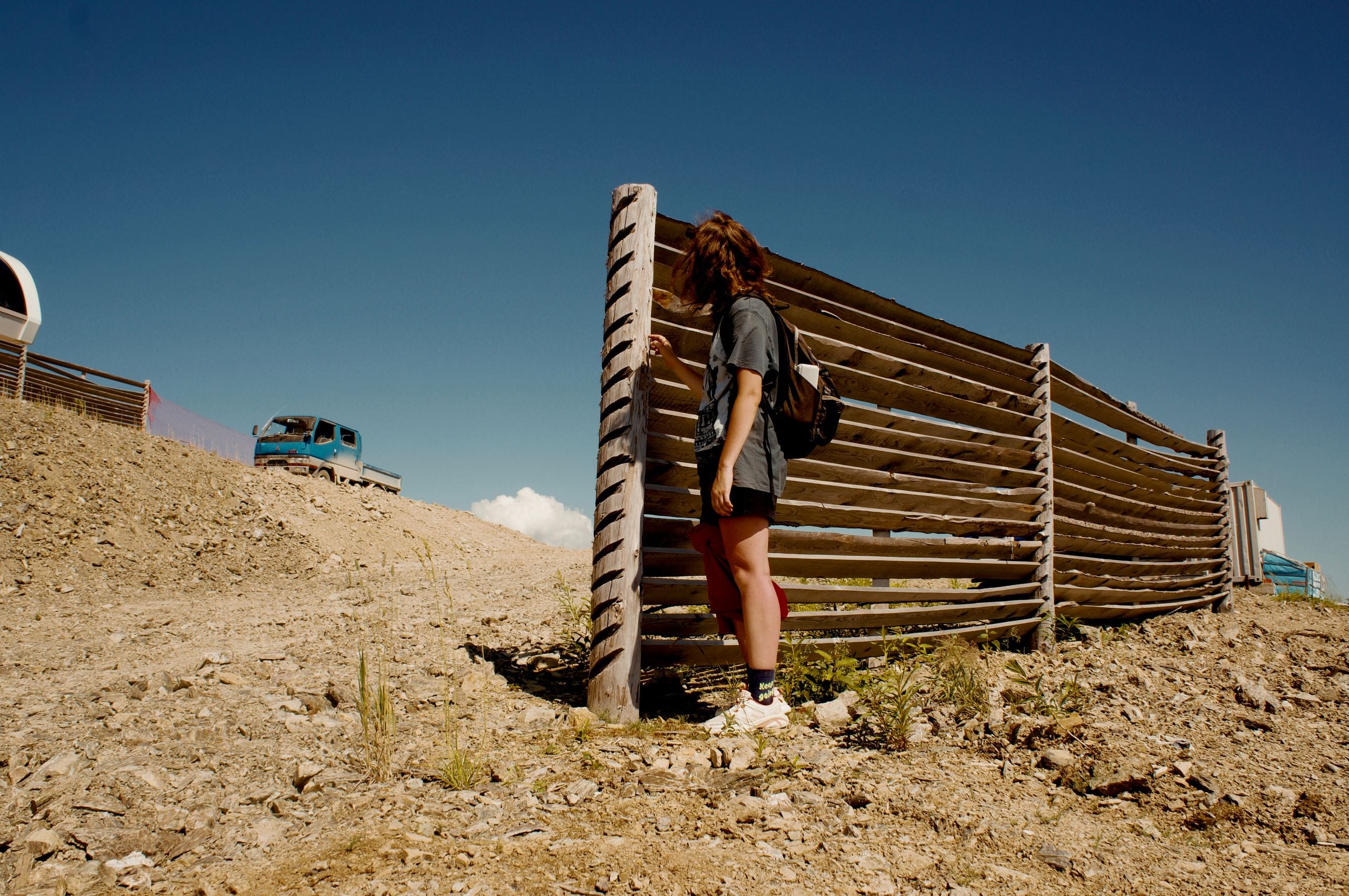 A woman stands by a wooden fence on a sunny day, with a truck in the background.